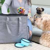 Women wearing grey sweater reaching into grey canvas and nylon bag, similar to backpack material. Bag is grey with side pockets, black zipper at top, and handle hanging on back. A pug and golden doodle are looking at the toy the woman is taking out of the bag. The bag is displayed with two collapsible blue dog bowls.
